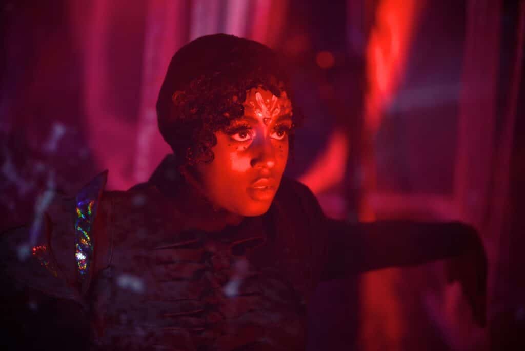 Nwando Ebizie looks forward with red light shining on her face, which has decorative gems and paint on it.