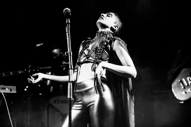 Black and white image of Juliana performing on stage, leaaning back with her hand on her hip and head pointing up up towards the ceiling